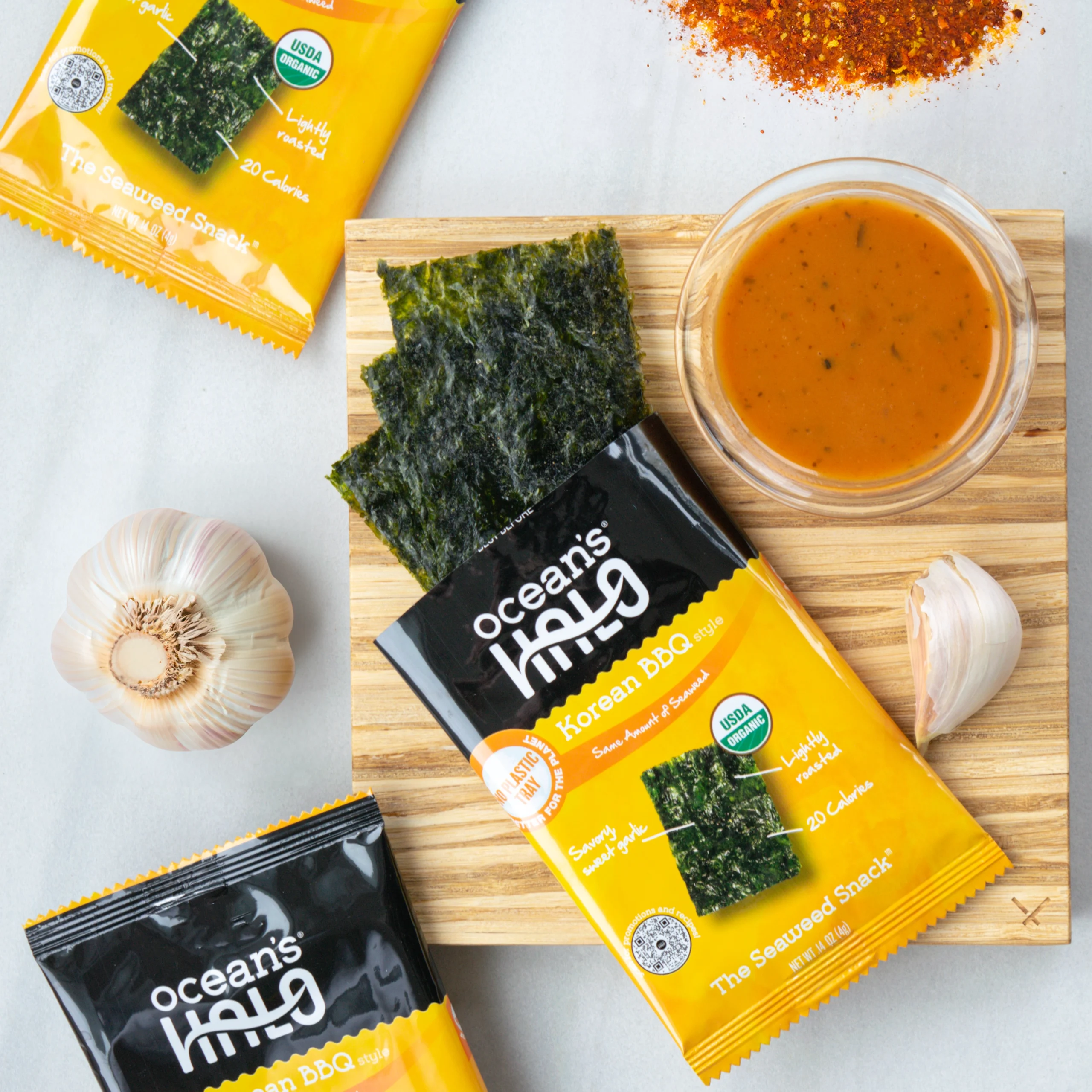 Ocean's Halo Korean BBQ seaweed snack packages on a wooden board next to garlic, chili powder, and dipping sauce.
