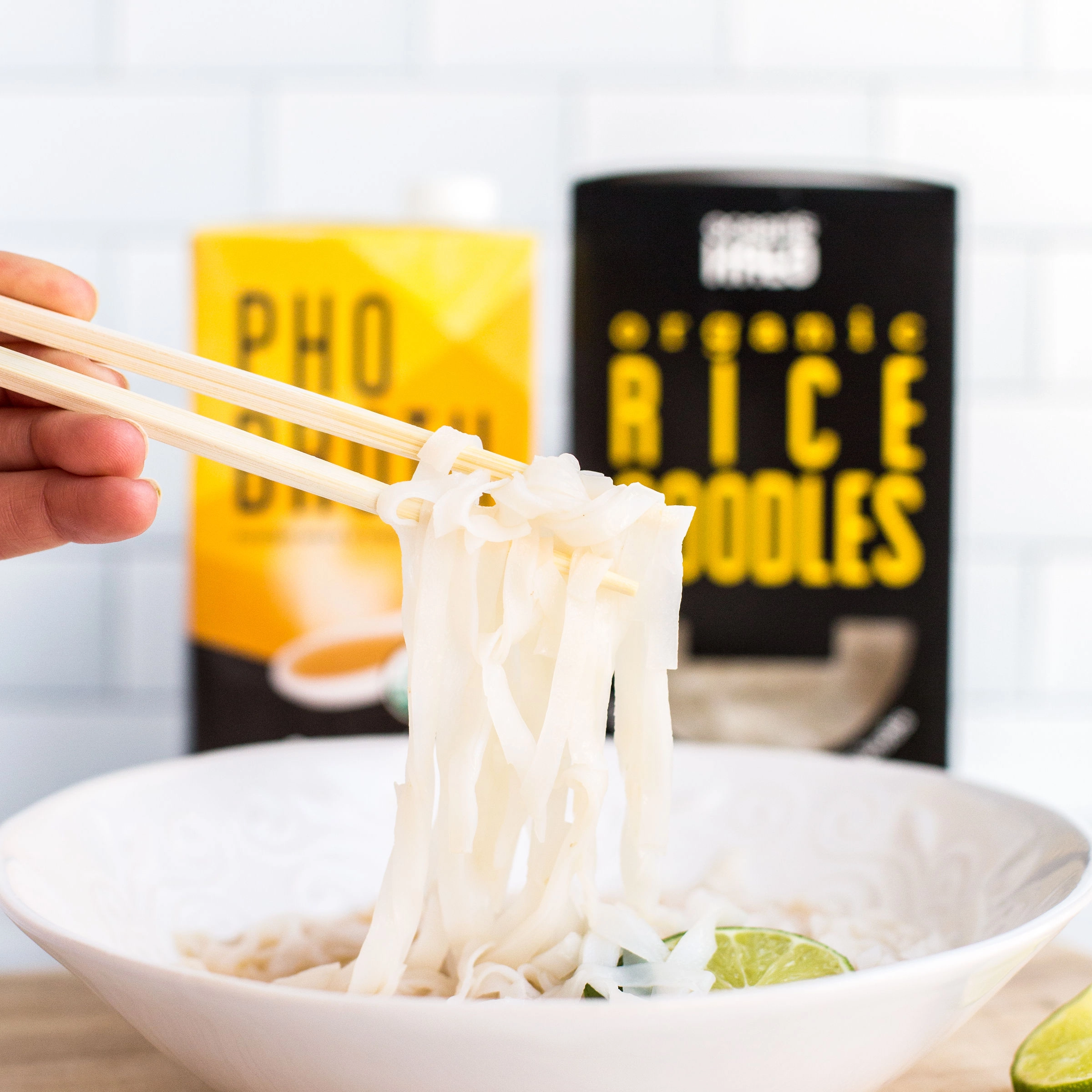 Chopsticks lifting rice noodles from a bowl in front of Ocean's Halo Pho Broth and Organic Rice Noodles packaging.