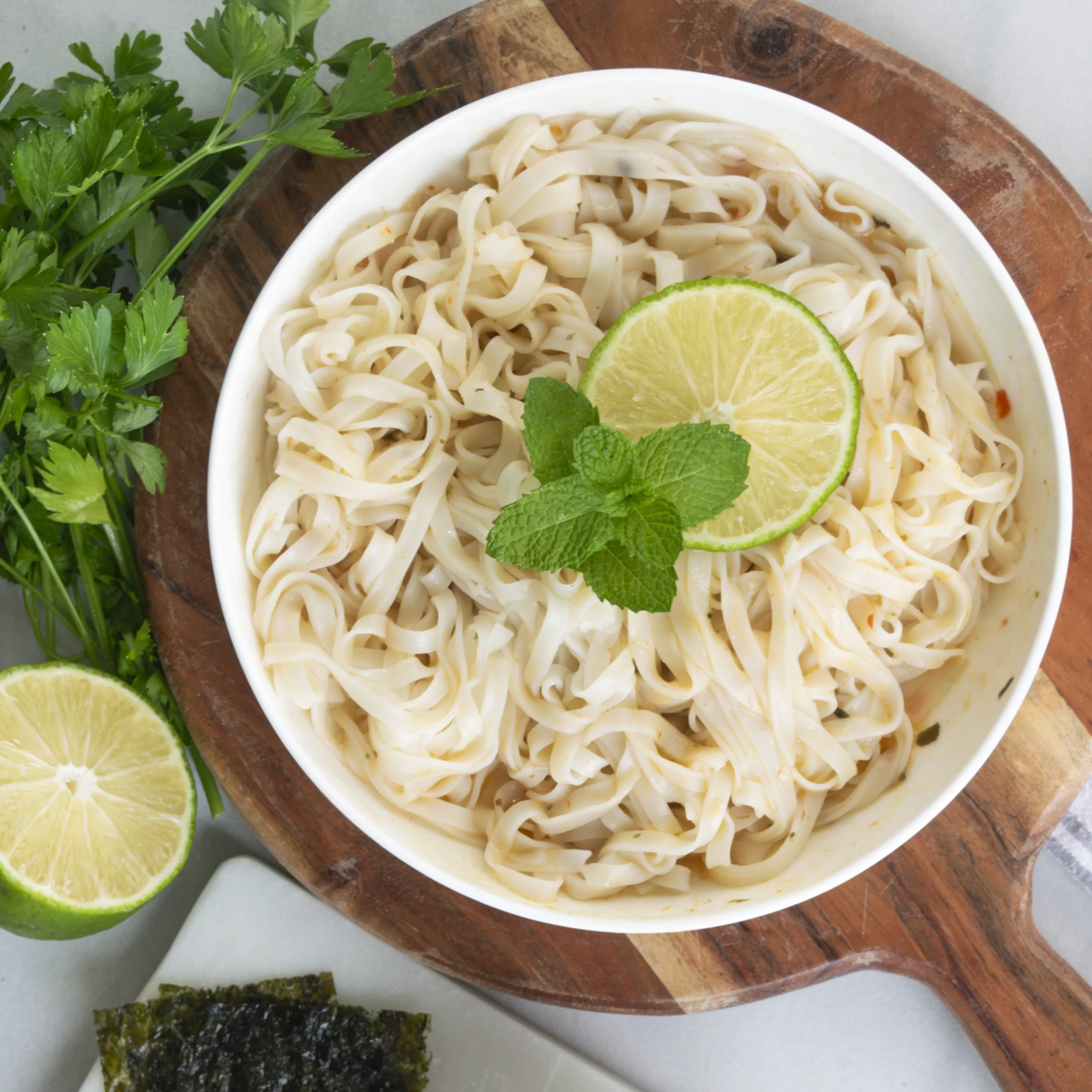 Top-down view of a bowl of cooked flat rice noodles garnished with a lime slice and fresh mint leaves.