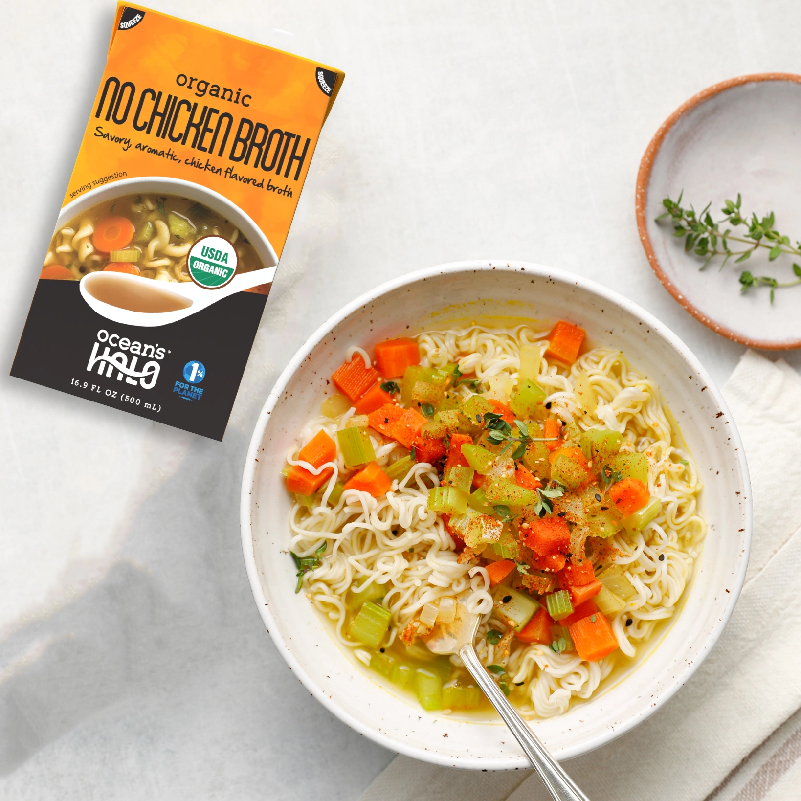 Bowl of noodle soup with carrots and celery next to Ocean's Halo No Chicken Broth carton.