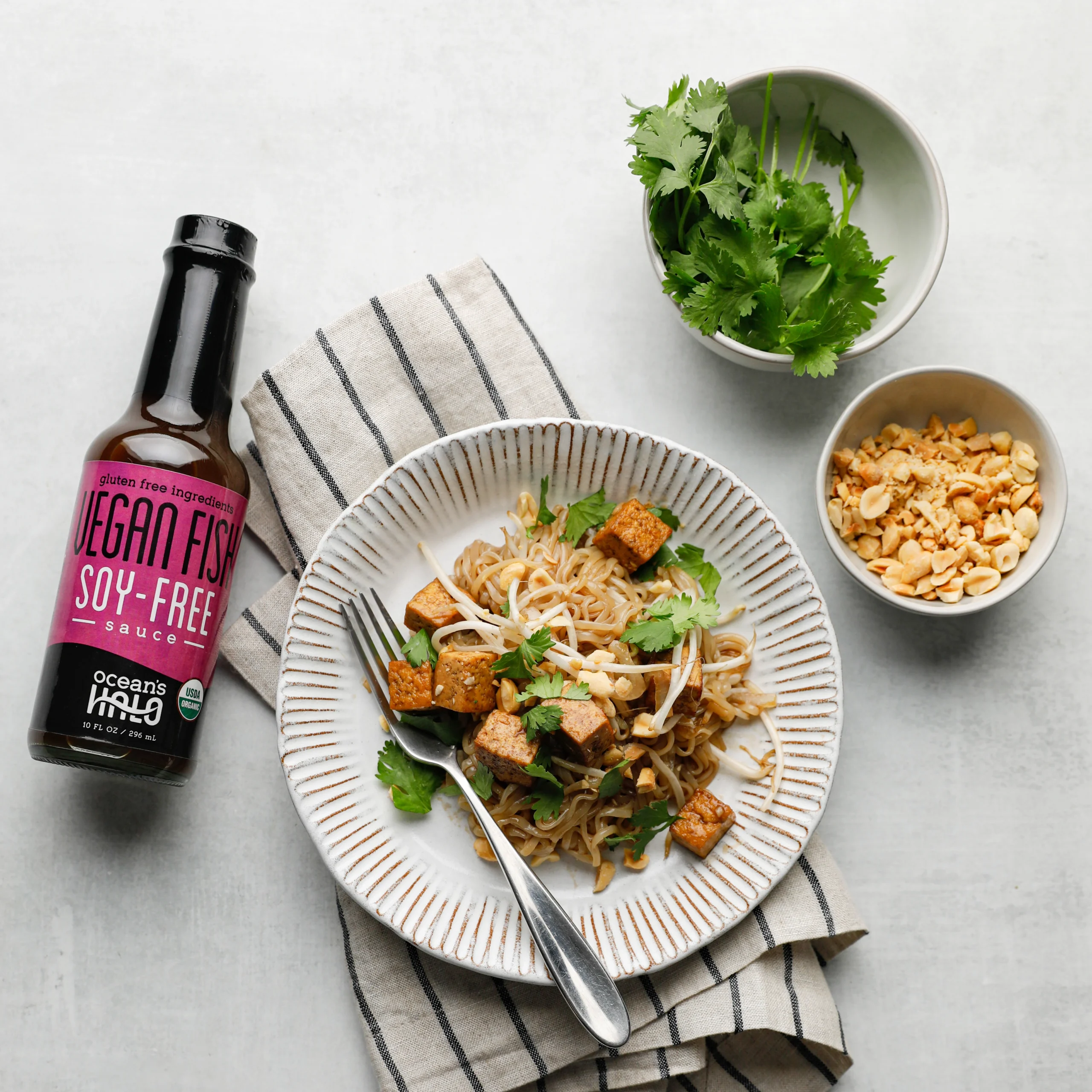 Tofu pad Thai noodles in a bowl garnished with cilantro next to a bottle of Ocean's Halo Vegan Fish Sauce.