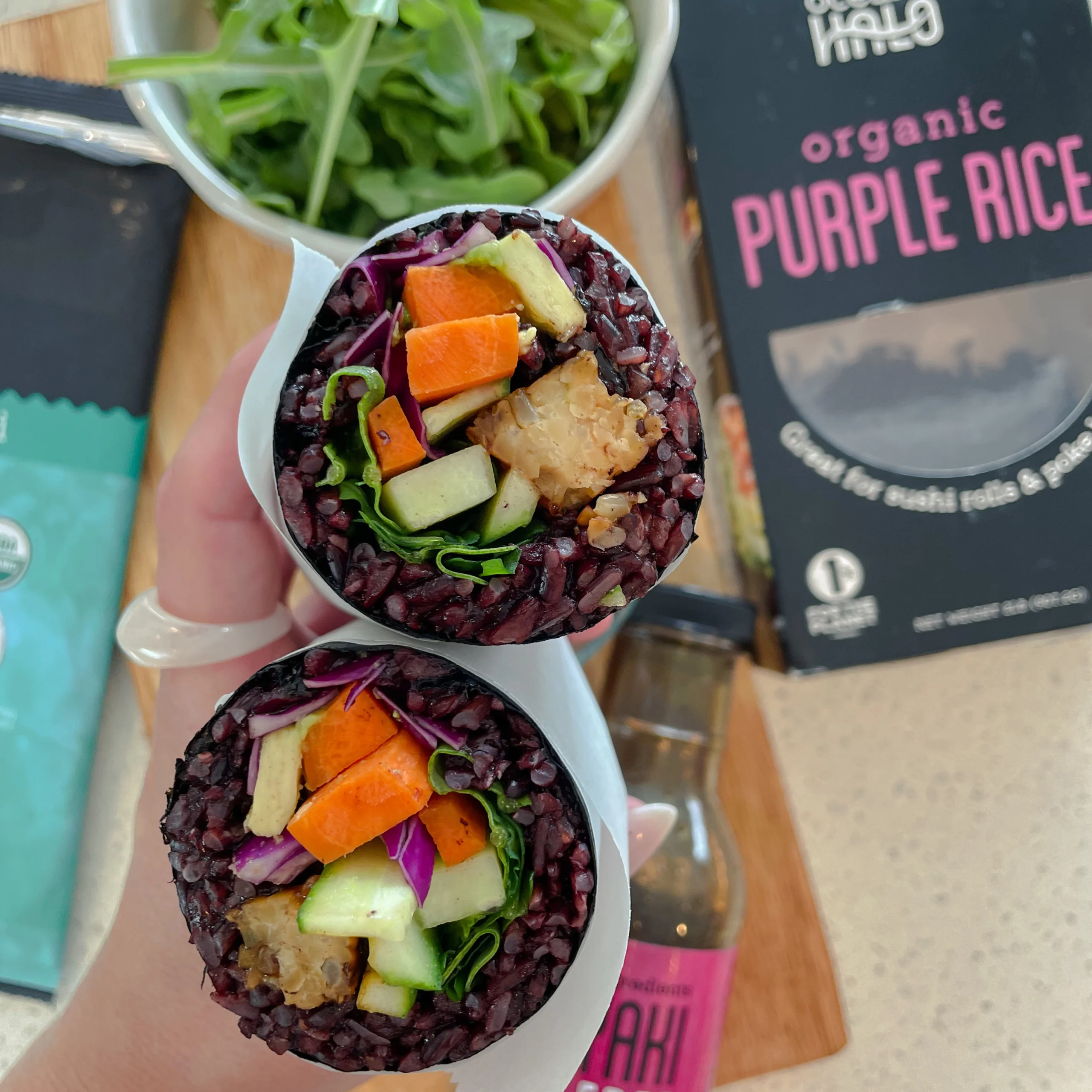 Hand holding a cut sushi burrito made with purple rice and fresh veggies next to Ocean's Halo Purple Rice packaging.