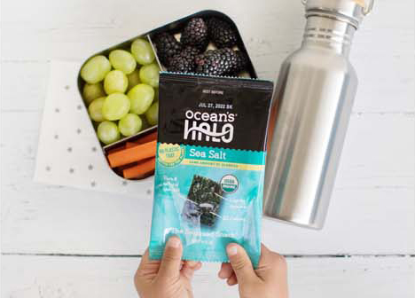 A kid holding Ocean’s Halo Sea Salt Seaweed Snacks over a lunchbox filled with grapes, carrots and blackberries.