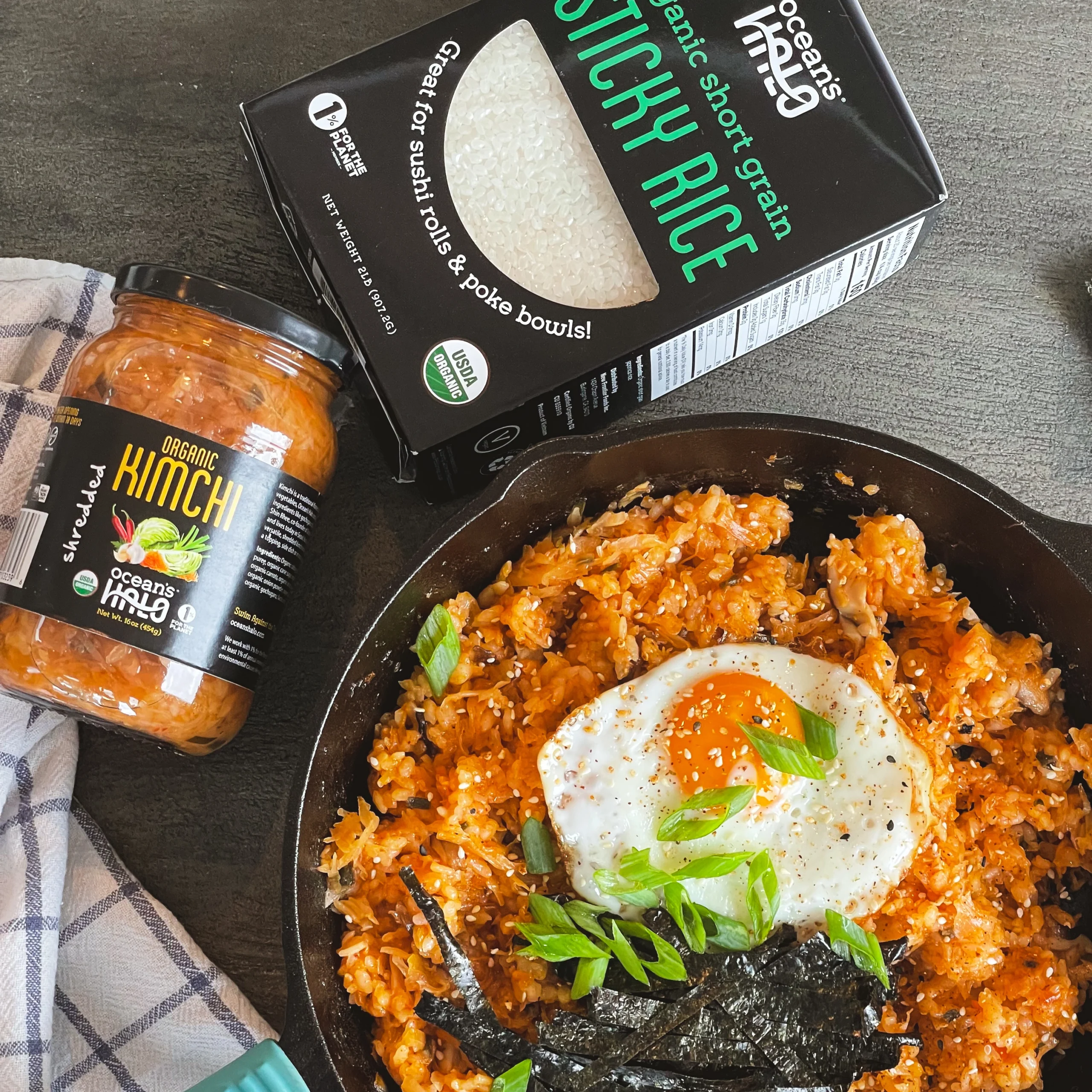 Kimchi fried rice topped with a sunny-side-up egg, sliced green onions, and seaweed strips in a black cast-iron skillet next to a jar of Ocean's Halo kimchi and a box of sticky rice.