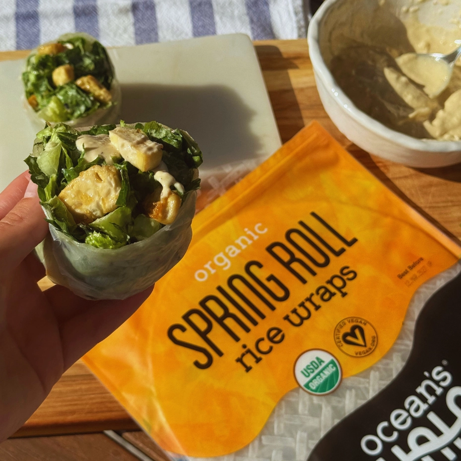 A hand holding a Caesar salad spring roll wrapped in an Ocean's Halo Rice Wrap, with packaging in the background