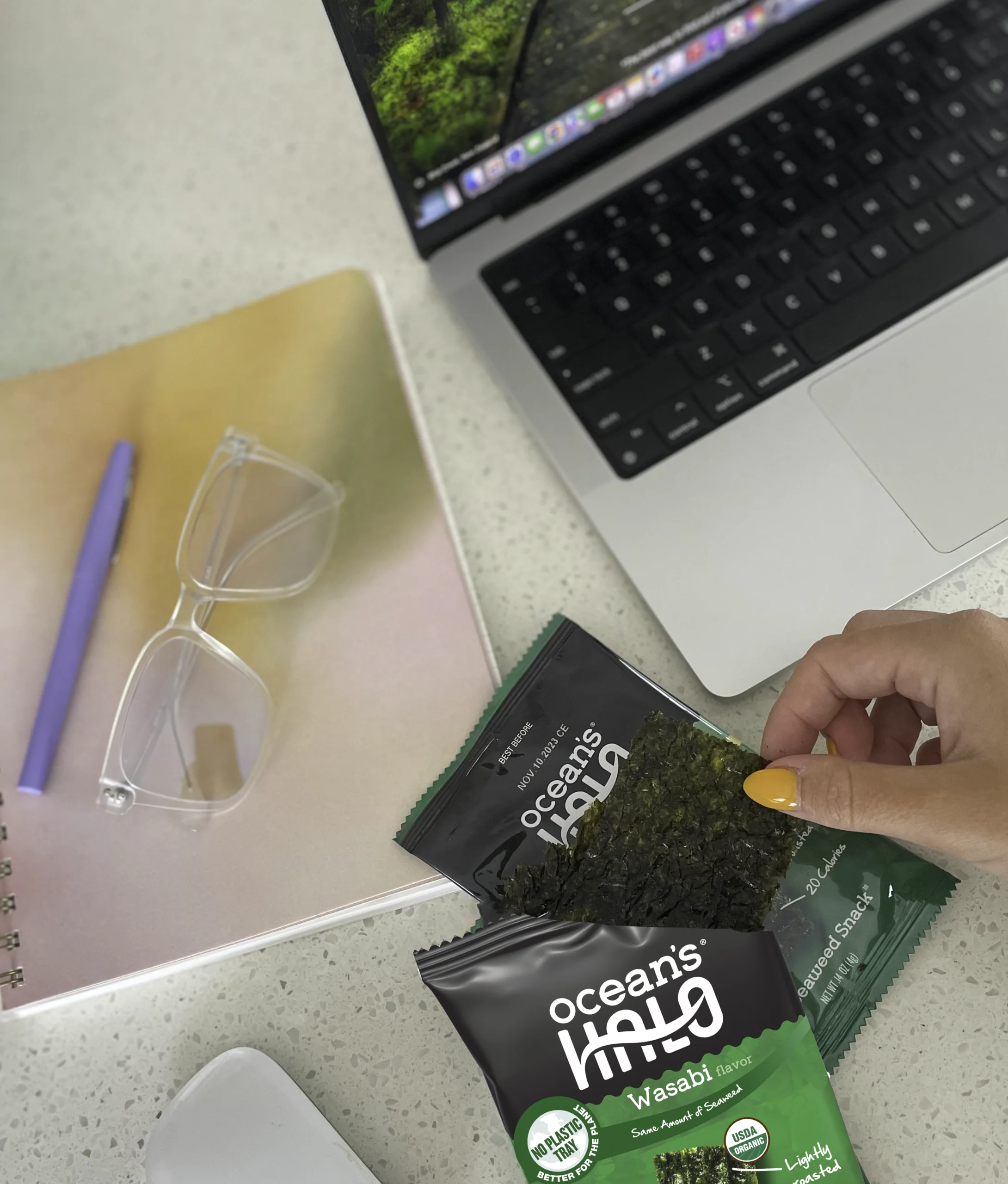 A hand holding a crisp seaweed snack over an Ocean's Halo Wasabi package at a desk next to a laptop and notebook.