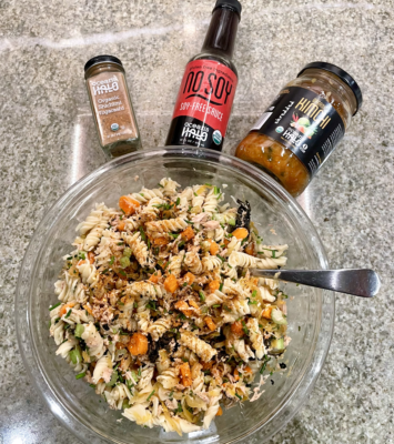 A bowl of pasta salad accompanied by a bottle of sauce and a jar of dressing on a wooden table.
