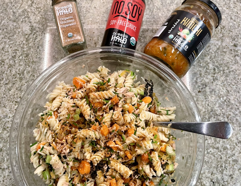 A bowl of pasta salad accompanied by a bottle of sauce and a jar of dressing on a wooden table.