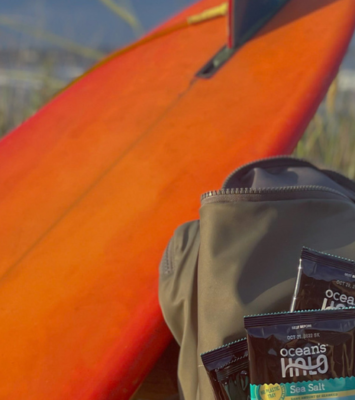 A backpack full of Ocean’s Halo Sea Salt Seaweed Snacks next to an orange surfboard in front of the beach & the ocean.
