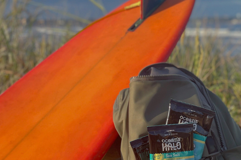 A backpack full of Ocean’s Halo Sea Salt Seaweed Snacks next to an orange surfboard in front of the beach & the ocean.