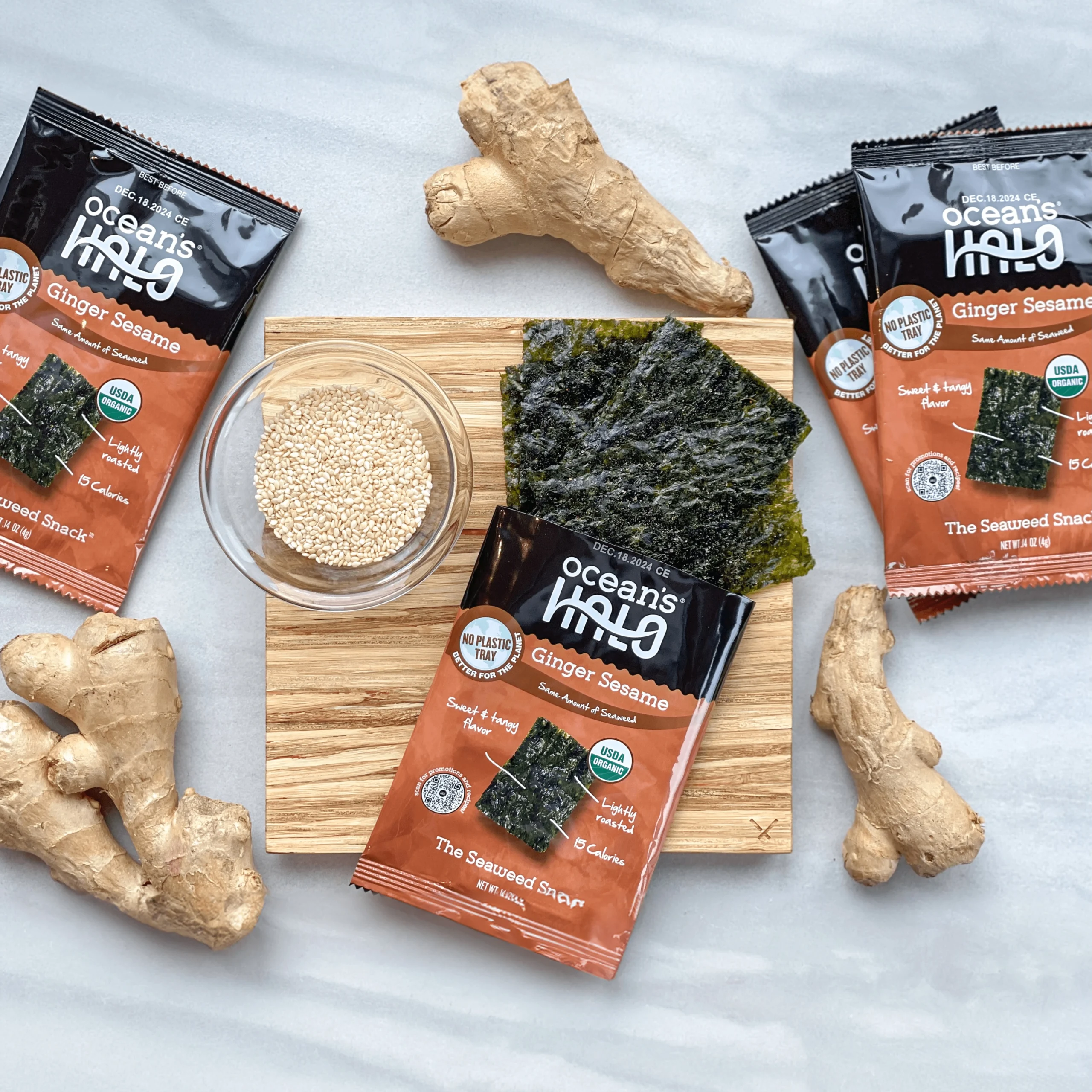 Ocean's Halo Ginger Sesame seaweed snack packages on a wooden board with raw ginger root and sesame seeds.