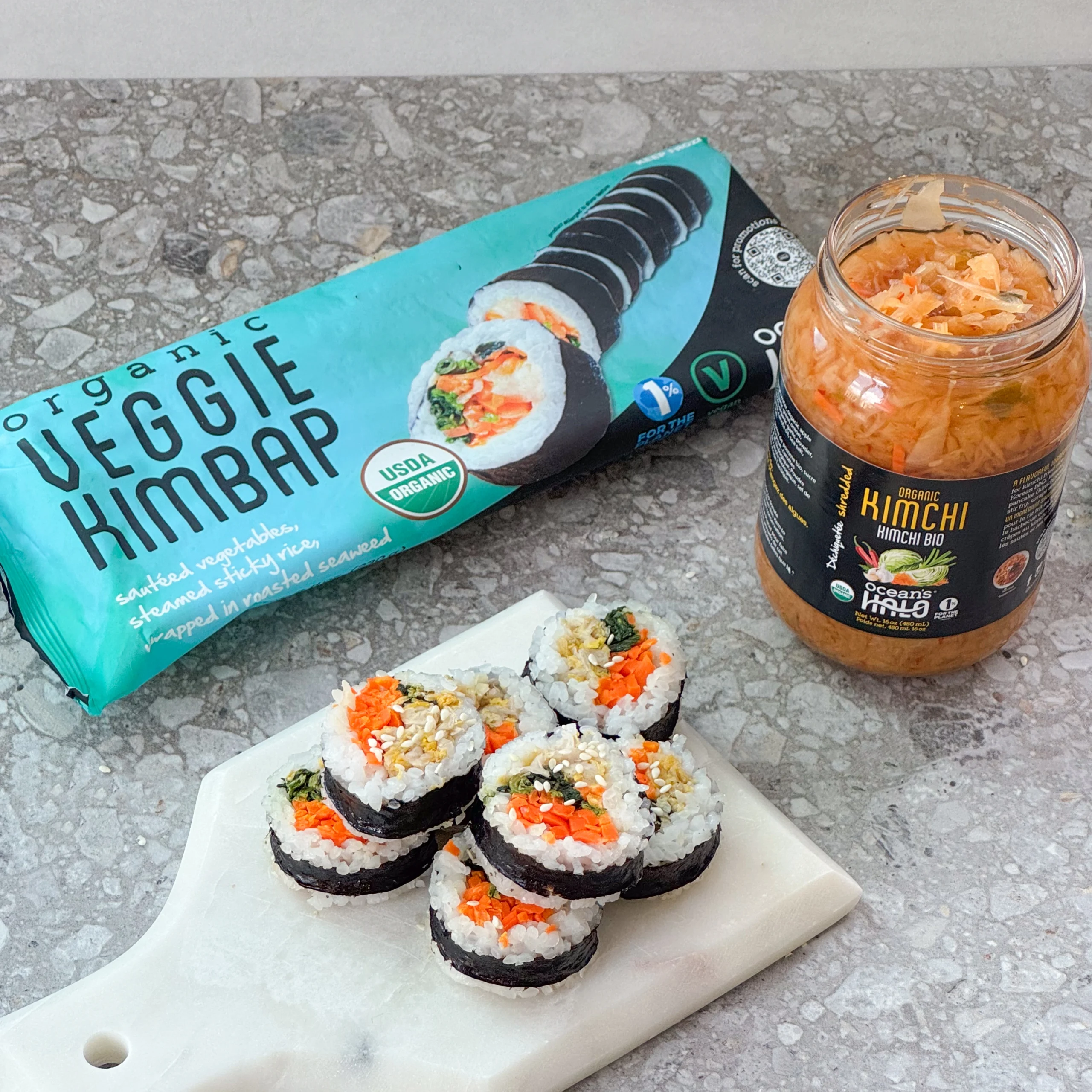 Sliced veggie kimbap on a white marble tray next to a jar of kimchi and Ocean's Halo Veggie Kimbap packaging.