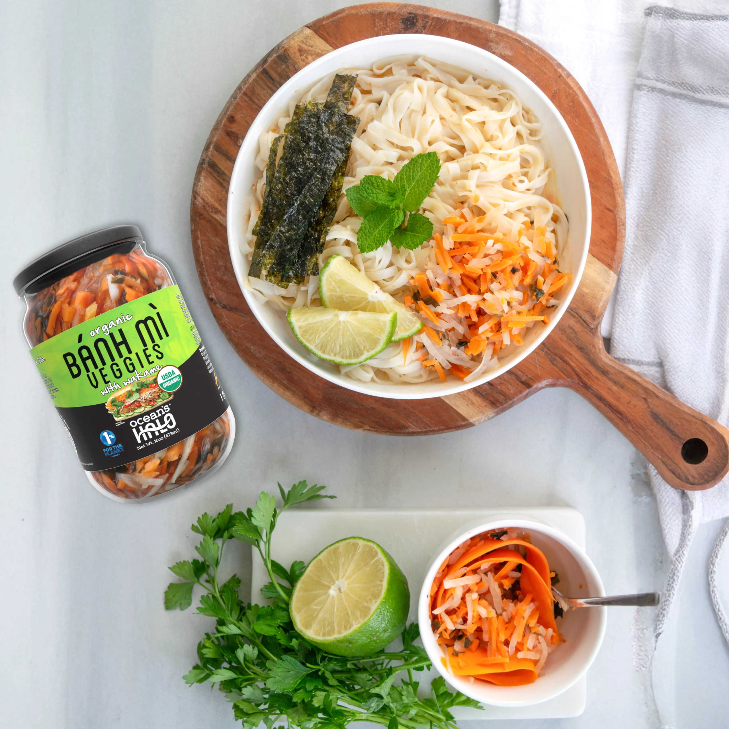 A noodle bowl topped with lime, mint, and pickled carrots next to a jar of Ocean's Halo Bánh Mì Veggies.