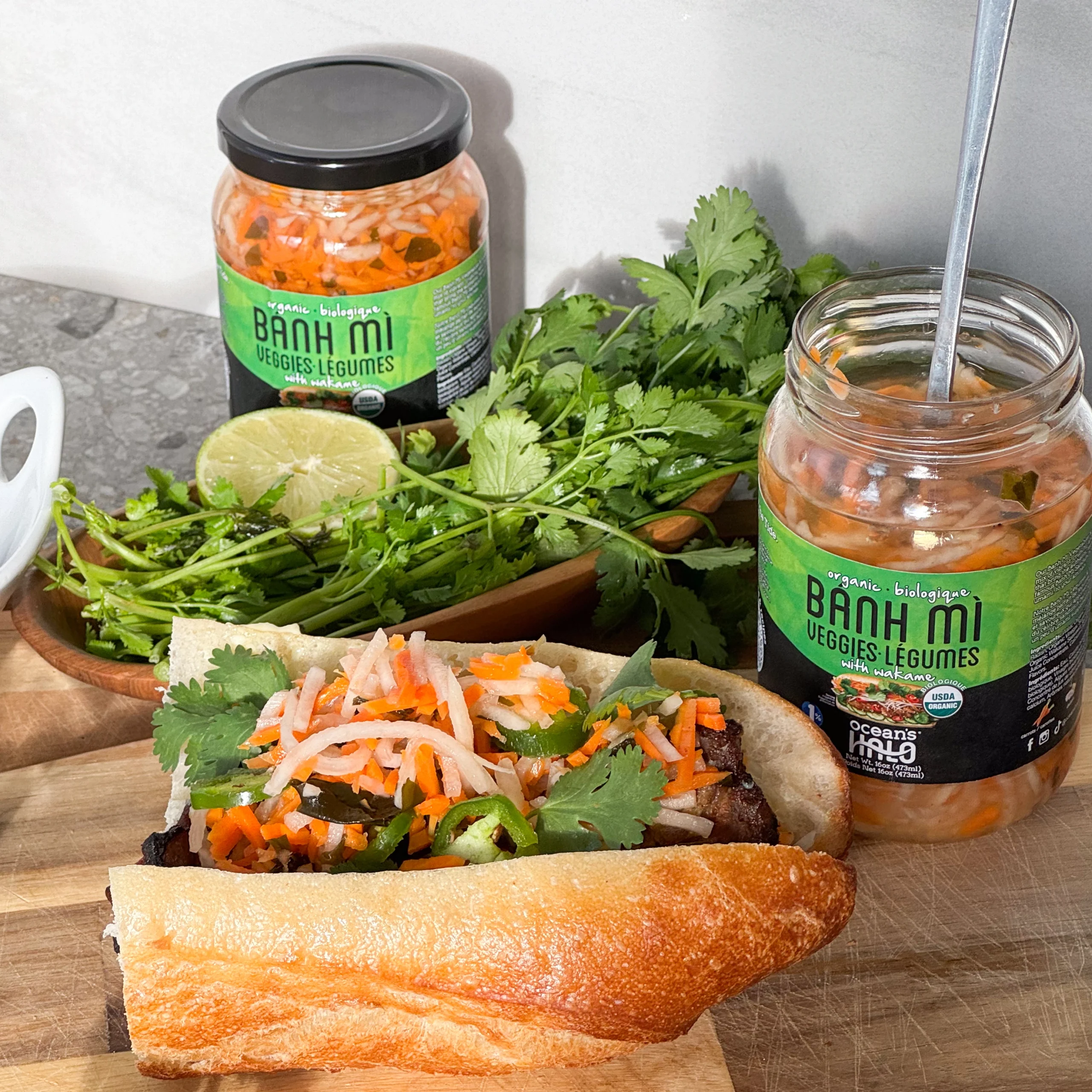 A fresh bánh mì sandwich stuffed with pickled veggies, cilantro, and meat next to jars of Bánh Mì Veggies