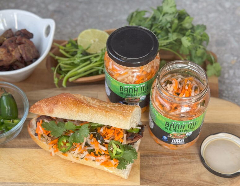 A banh mi sandwich next to jars of Ocean's Halo Banh Mi pickled vegetables.