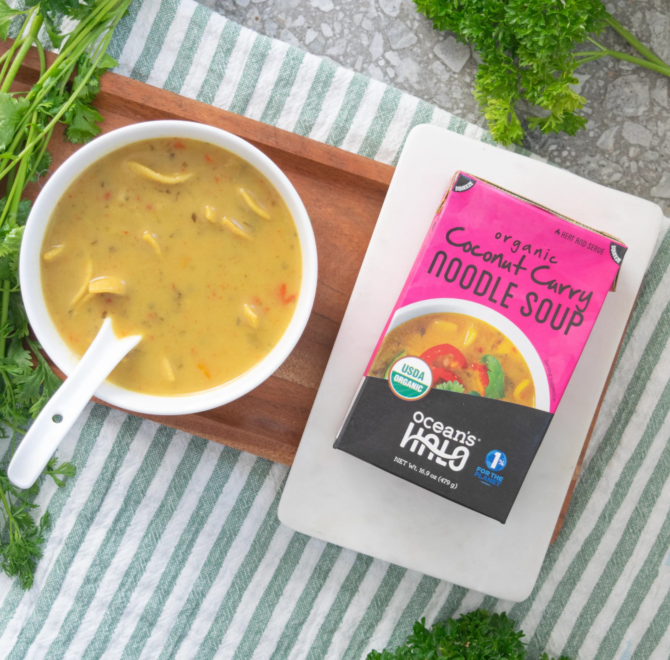 A flat-lay photo with a white bowl of noodle soup and the product box, resting on wooden and white marble trays, surrounded by fresh cilantro on a striped cloth.