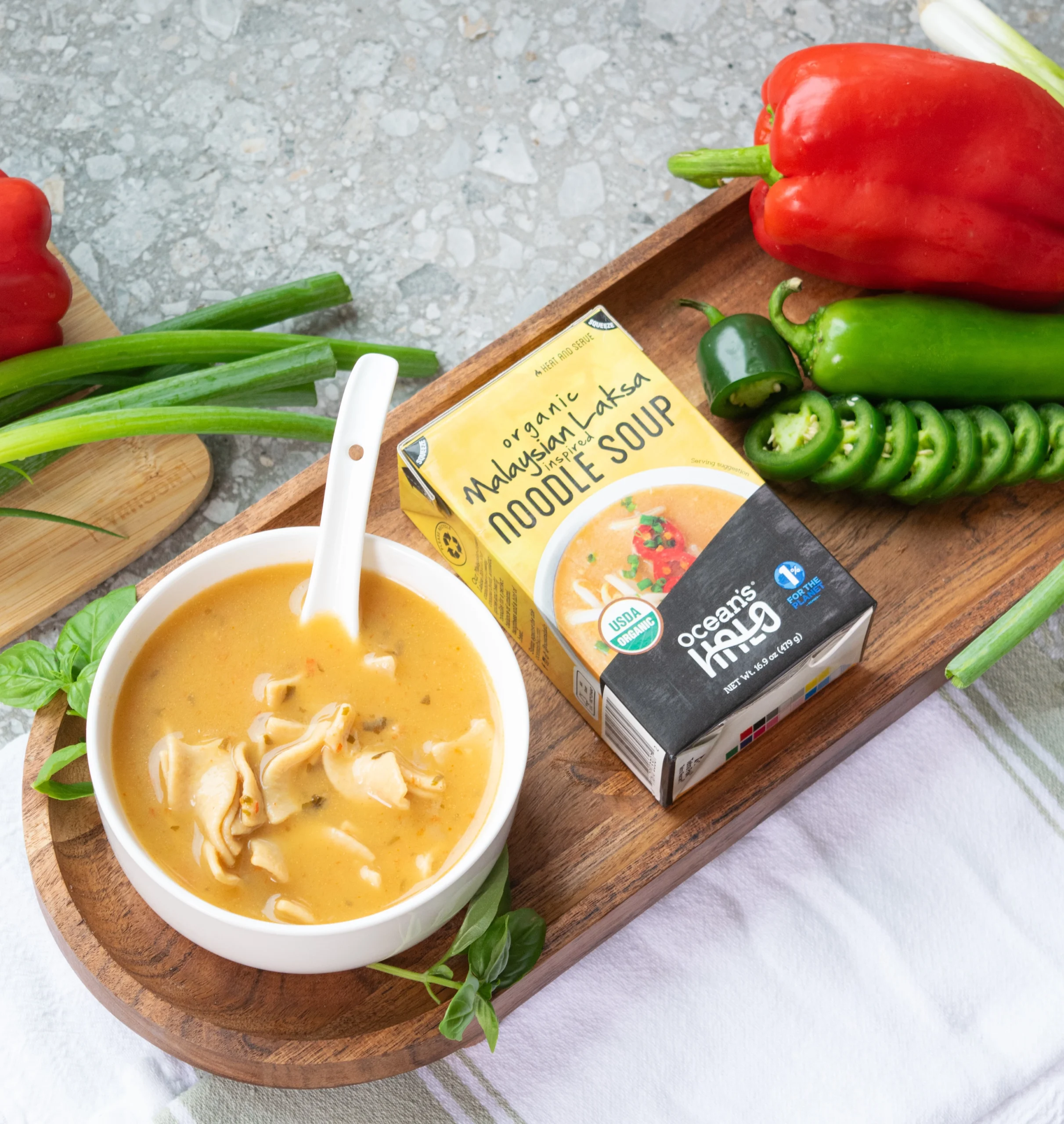 A bowl of noodle soup next to a carton of Ocean's Halo Organic Malaysian Laksa Inspired Noodle Soup on a wooden tray, surrounded by fresh ingredients like bell peppers, jalapeños, and green onions.