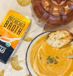 A bowl of creamy pumpkin soup with toast and seeds sits beside a carton of vegan "No Chicken Broth" on a fall placemat.