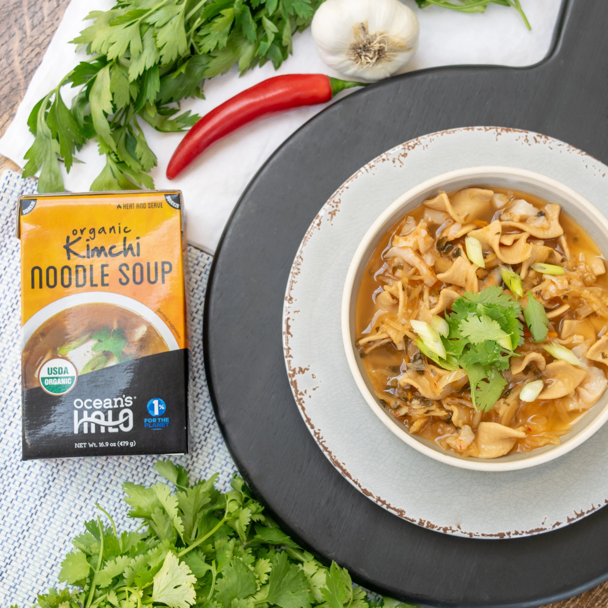 Top-down lifestyle view of a bowl of Kimchi Noodle Soup next to the carton, surrounded by fresh herbs, garlic, and a red chili pepper on a black serving board.