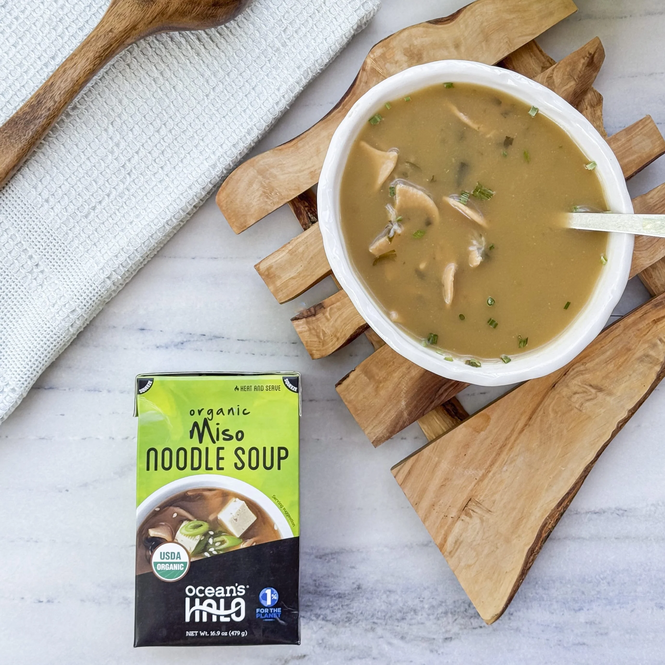 Top-down view of a bowl of Miso Noodle Soup served on a wooden trivet alongside the product carton, a wooden spoon, and a linen towel.