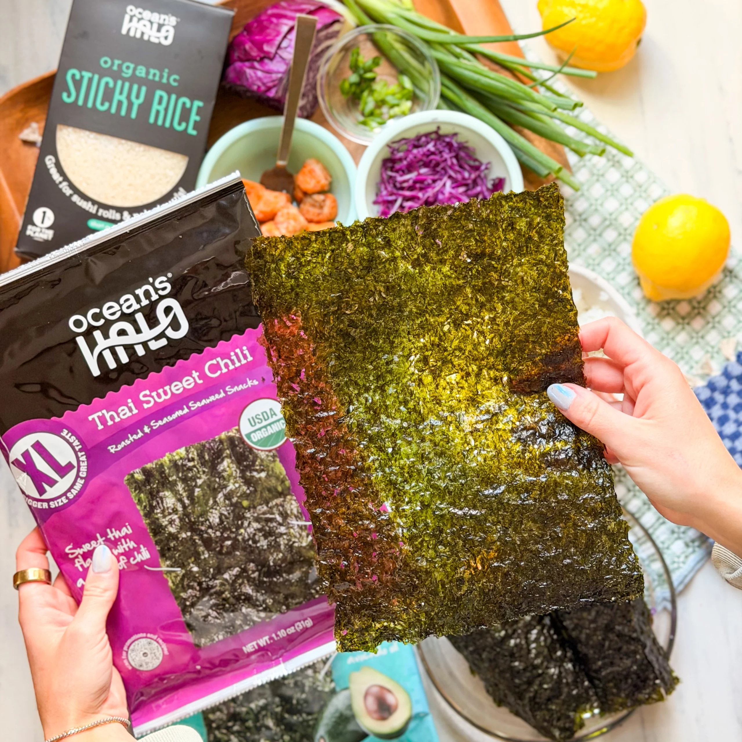 Hands holding a large seaweed sheet next to a purple Ocean's Halo XL Thai Sweet Chili package and fresh ingredients