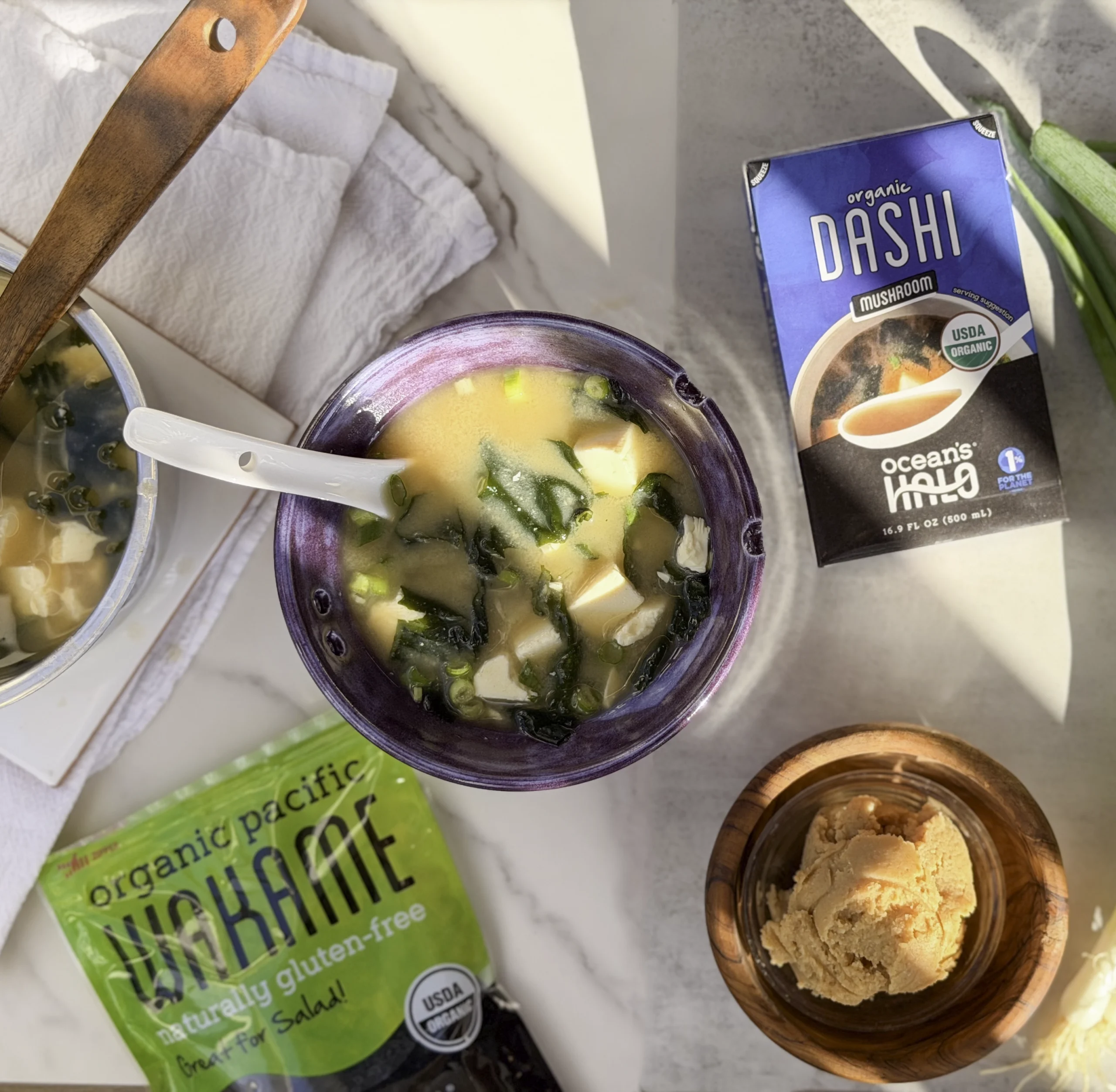 Miso soup image with ocean's halo wakame and mushroom dashi laying next to it. Miso paste and green onions in the scene.