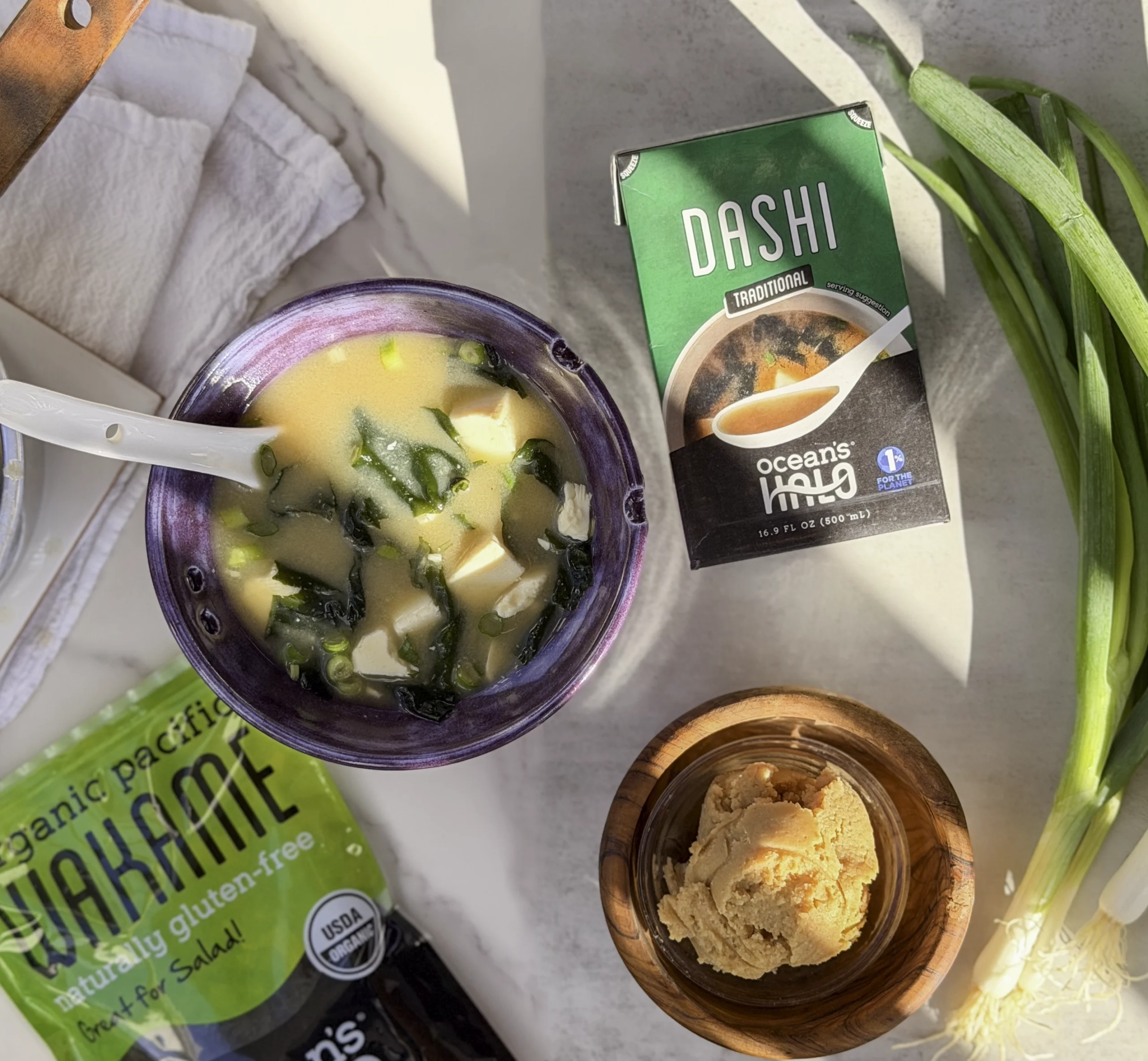 Miso soup image with ocean's halo wakame and traditional dashi laying next to it. Miso paste and green onions in the scene.