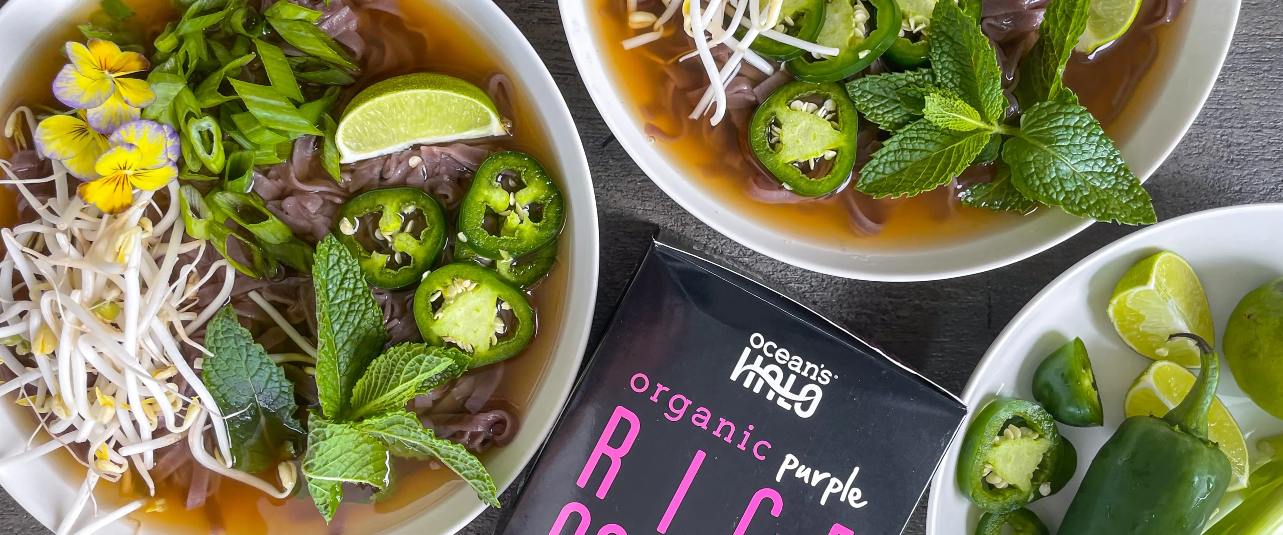 Two bowls of pho with purple rice noodles, limes, jalapeños, and sprouts next to Ocean's Halo Purple Rice Noodles.